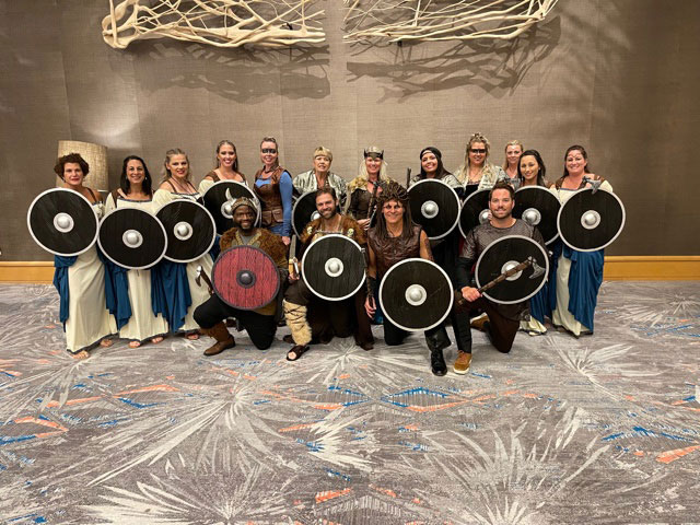 A group of people dressed in historical costumes, posing with Viking-style shields and weapons, standing on a stage with a backdrop featuring a large wooden frame.