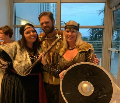 The image shows three individuals dressed in costumes resembling historical warriors, posing together with one person holding a shield and another wearing a helmet, standing indoors with a window in the background.