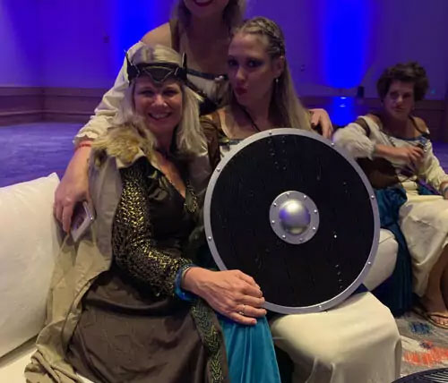 The image shows two individuals dressed in costumes with Viking-style elements, posing with a large shield, and they are seated on a couch, with one person holding the shield and smiling at the camera.
