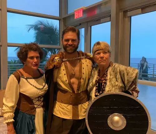 Three people dressed in medieval costumes standing together with a backdrop of ocean and sky.